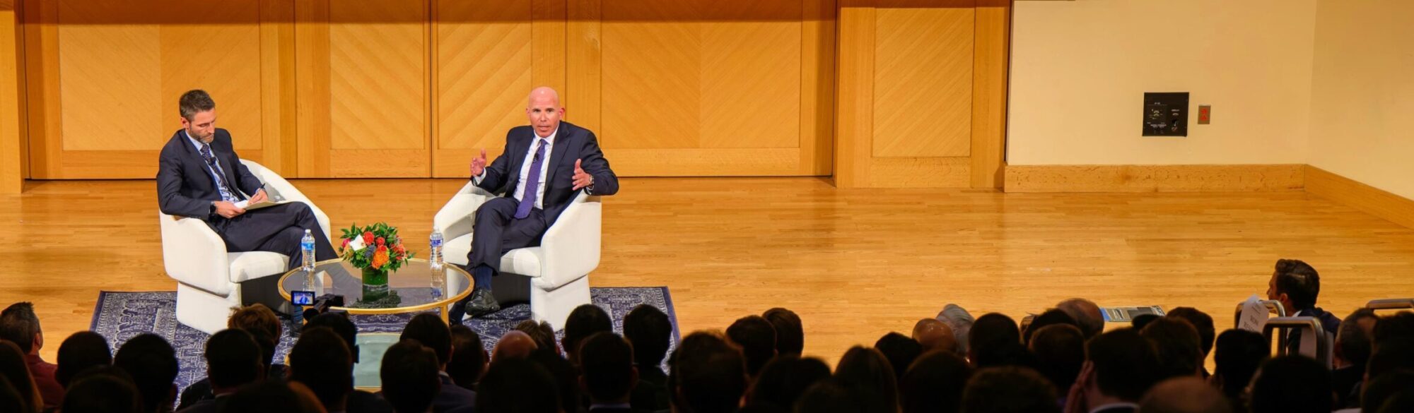 2024 Luminaries Main Speaker, Scott Rechler speaking to an audience along with Director, Matthew Cypher as moderator, both are chatting in armchairs at the center of a stage. Water bottles and floral decorations on top of a glass table in front of them. Stage is well-lit while lighting in audience is dimmed to illuminate the stage.