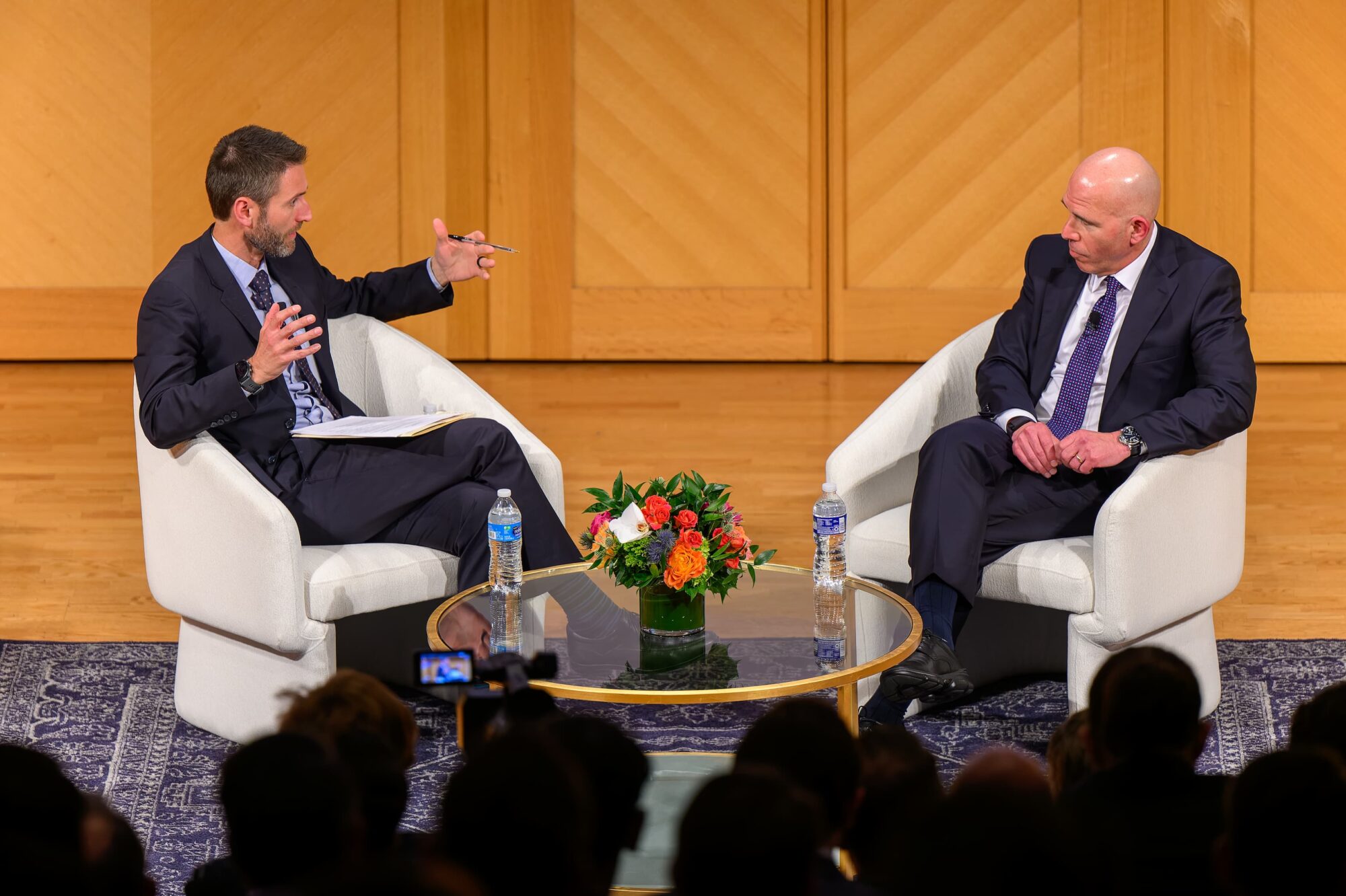 Director, Matthew Cypher (as moderator) speaking to 2024 Luminaries main speaker, Scott Rechler. Both are sitting in chairs in front of an audience. Water bottles and floral decorations on top of a glass table in front of them.
