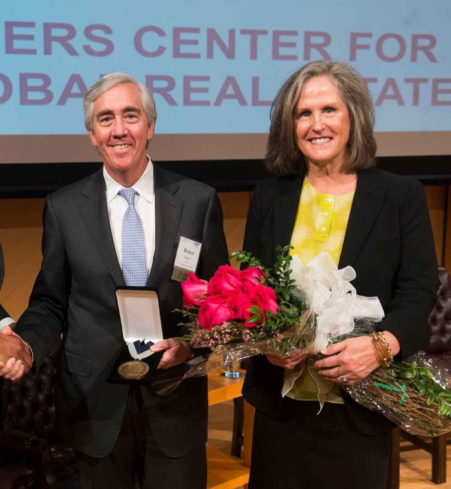 Bob Steers and Lauren Steers posed with the former dean, David Thomas. Lauren Steers holds flowers, and Bob Steers is holding a medal of appreciation and shakin David Thomas's hand.