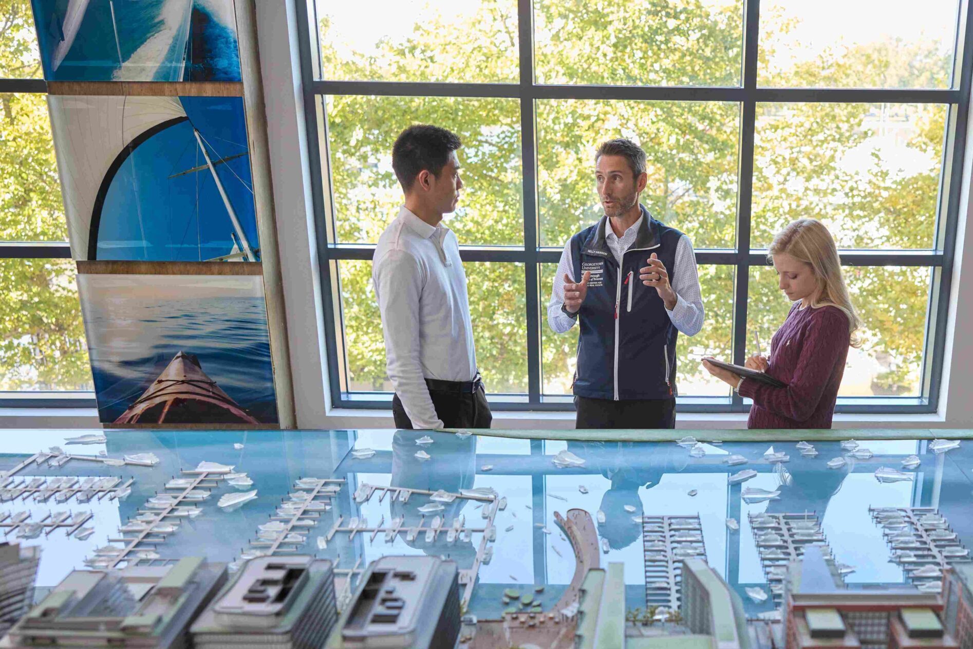 Director, Matthew Cypher is speaking to two Steers students while standing around a detailed architectural model of a waterfront development. Large windows in the background let in natural light, illuminating the model and the group as they discuss.
