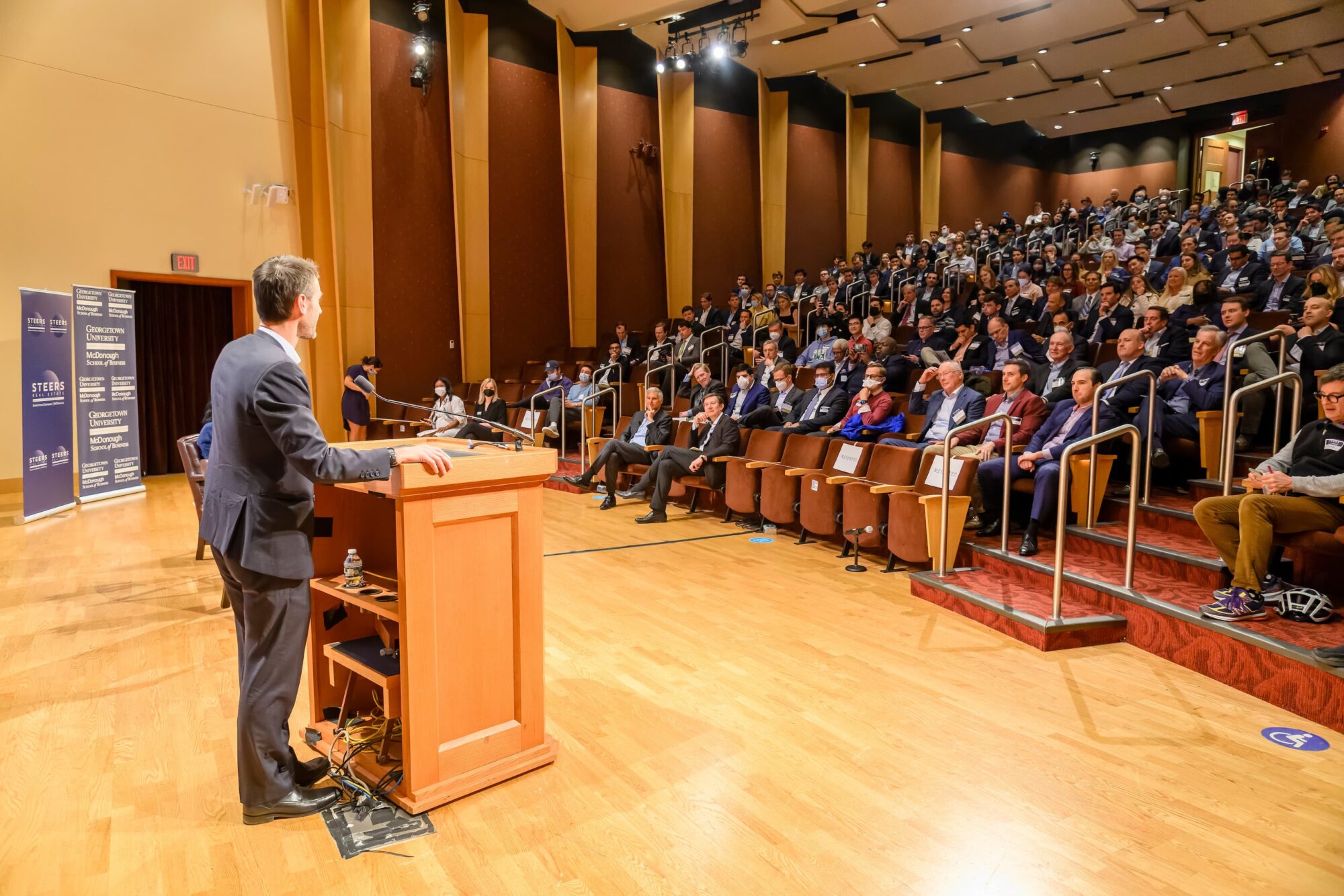 A speaker stands at a podium, addressing a large audience seated in an auditorium. The room is well-lit, with people attentively listening. The audience includes individuals of various ages, dressed primarily in formal attire.