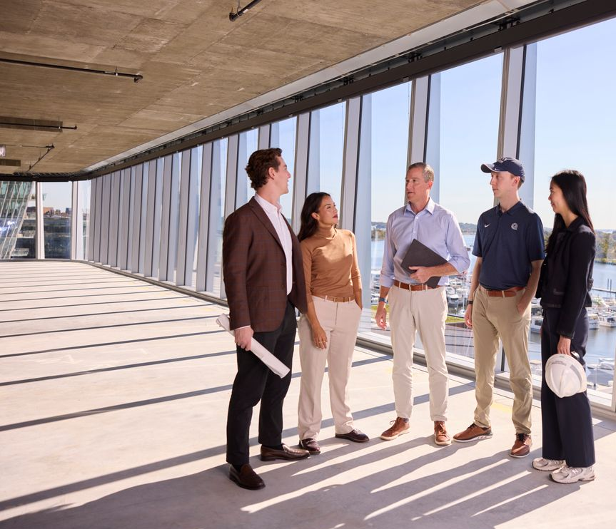 Executive Director, Tim Reardon (middle) speaking with Steers real estate students in business casual attire in a building overlooking a waterfront. Large windows letting in light that is illuminating the group on a clear and sunny day.