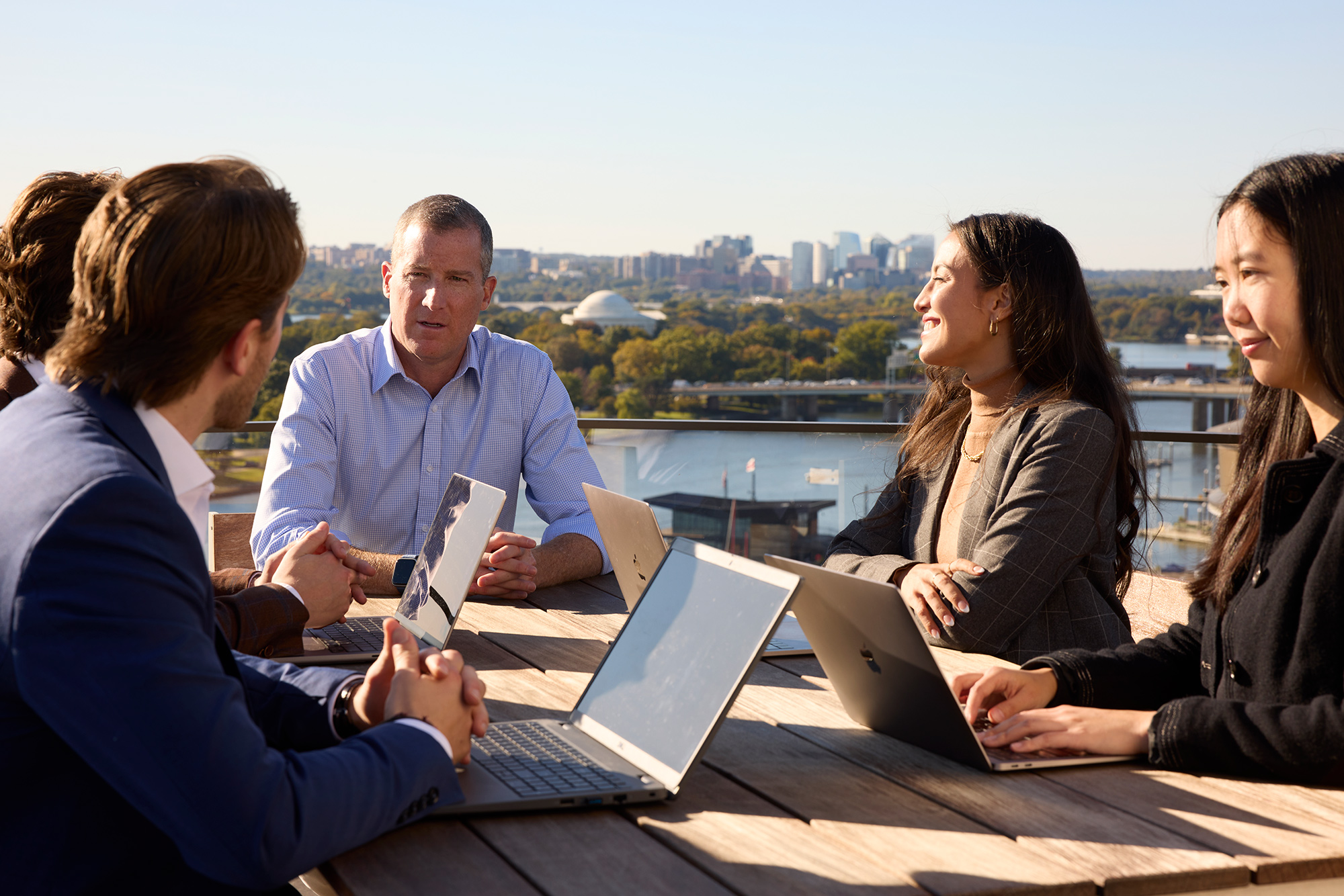 Executive Director, Tim Reardon sitting with four students at a table outside, chatting outside with a cityscape in the background.
