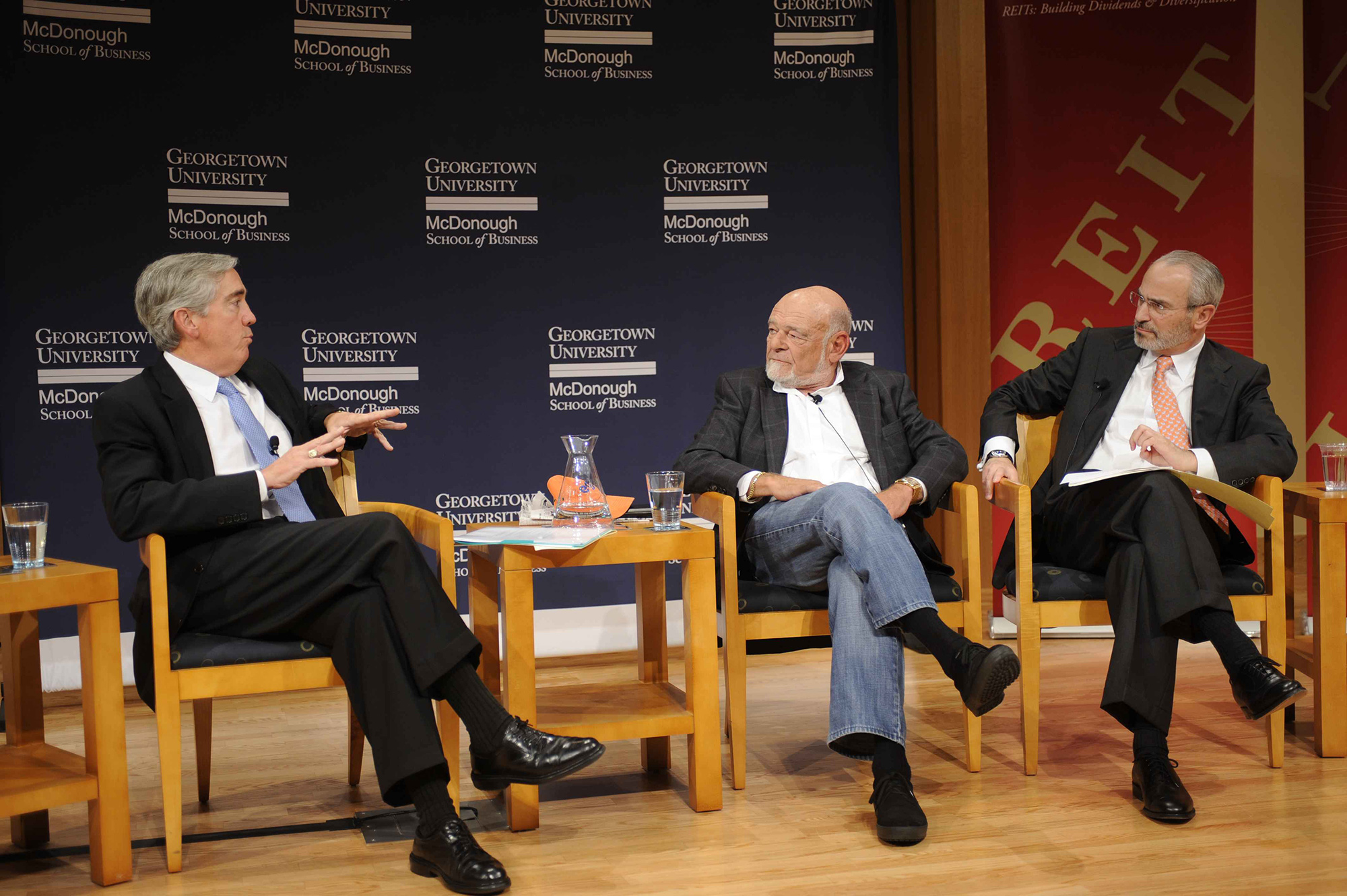 Steer Center Founder, Robert Steers, along with two guest speakers, all in business formal attire, engaged in a discussion on a stage with Georgetown University signage in the background. They are seated in wooden chairs, with books and cups of water on the table between them.