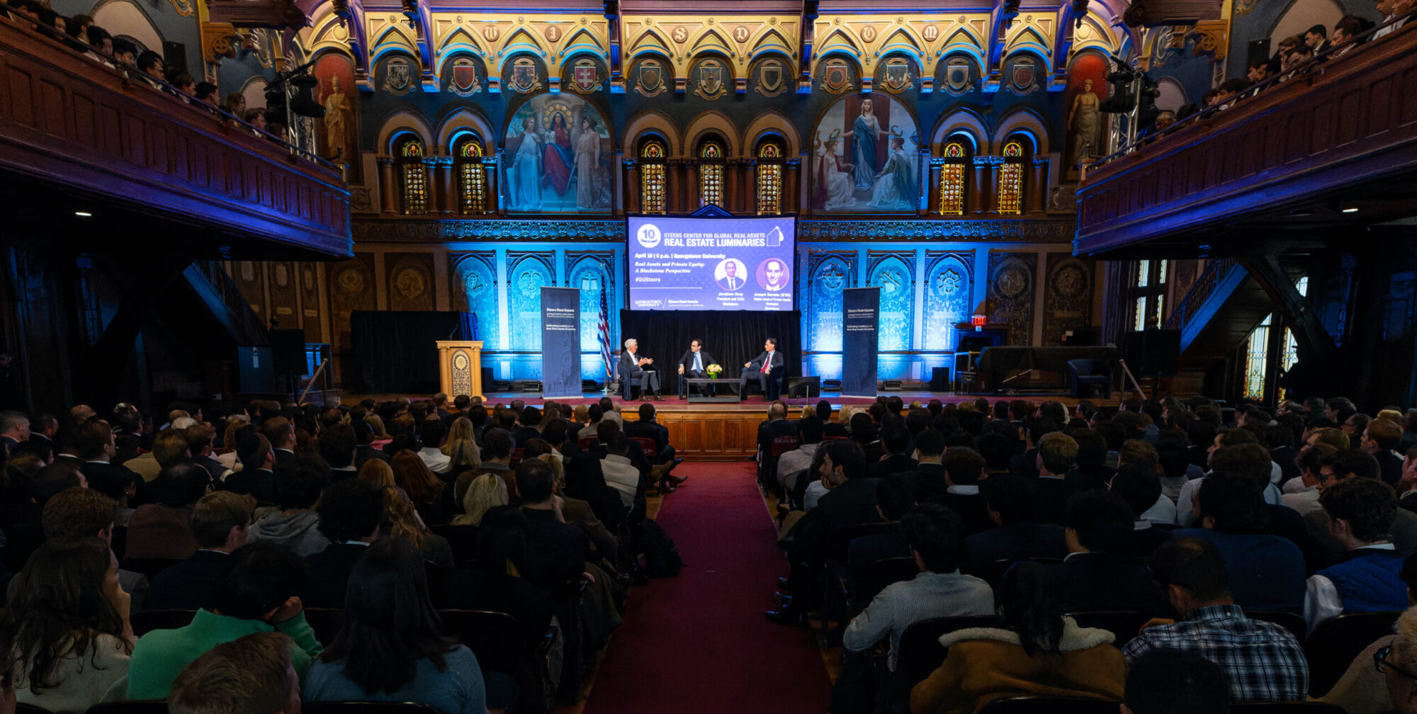 Luminaries 2025 photo in Gaston Hall of audience looking at illuminated main stage where Robert Steers, Joseph Baratta and CEO of Blackstone, Jonathan Gray. All three on the main stage are sitting on chairs in a semi circle with the Luminaries 2025 flyer on a projected slide.