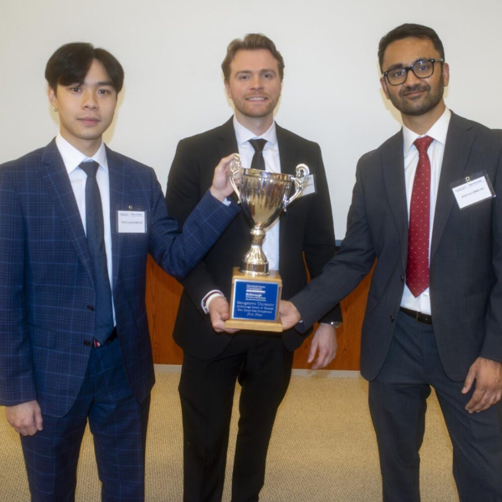 Steers real assets students who placed 1st in the 2025 McDonough Real Assets Case Competition at the McDonough School of Business. One student in the middle is holding a trophy for winning a real estate case competition. All are in business casual attire in a case room. Adequately lit space with overhead lighting.